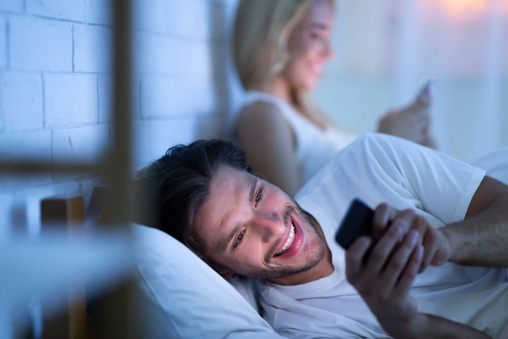 Nevera. Muž si v posteli píše s inou ženou